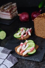 Two sandwiches with salted lard strips with meat strips and fresh cucumber on rye bread on a slate board on a black concrete background. Recipes for sandwiches, snacks.