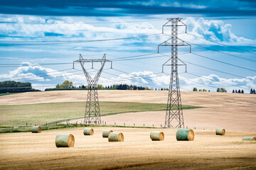 Transmission towers and power lines overlooking a farm field during fall harvest with round hay bales on the Canadian prairies in Rocky View County Alberta Canada.