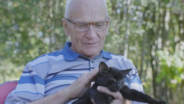 Grandfather seated in armchair on backyard and strokes black little cat on lap.