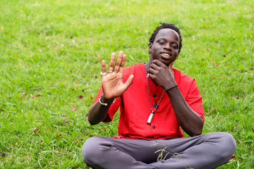 Young black man in red shirt sitting on the grass in a park waving.