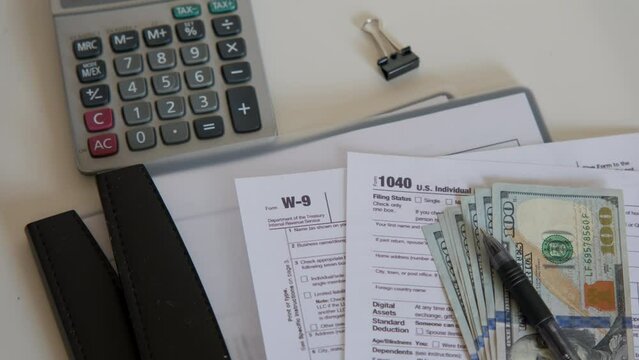 An American dollar tax bill, pen calculator and U.S. money. 1040 and W9 individual tax form are placed on desk of American citizen.