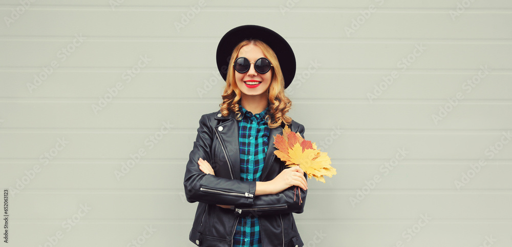 Wall mural Autumn portrait of stylish happy smiling young woman with yellow maple leaves wearing round hat and rock style black leather jacket posing on gray background