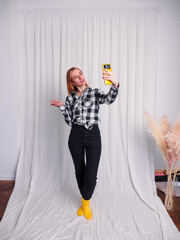 Cheerful blonde woman holds modern smartphone wears trendy stylish expresses happiness and joy models indoor. Studio shot selfie vlogger