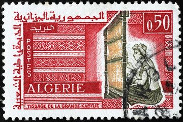 Woman creating a carpet at the loom on algerian stamp