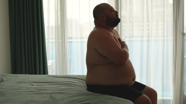 Fat middle aged man does exercises for breath to cope with summer stuffiness. Overweight obese chubby young male sitting on bed, difficult breathing deeply to handle with terrible heat, holding chest