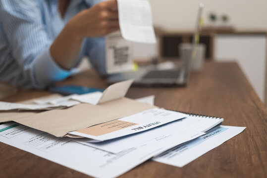 Utility bills with total amount for online payment through laptop lie on table wooden surface against woman examining bank documents in modern premise at home