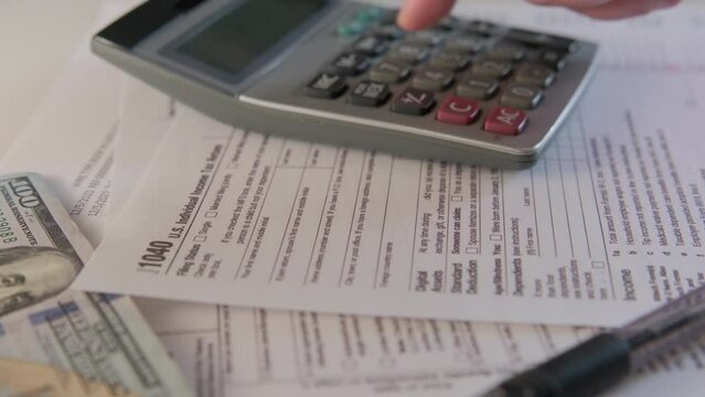 Close up shot of calculate the amount on a calculator 1040 and W9 individual tax form are placed on desk of American citizen. Move camera