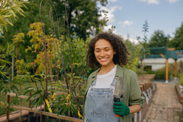 Naklejka premium Female gardener standing with hand trowel and looks camera on plants background