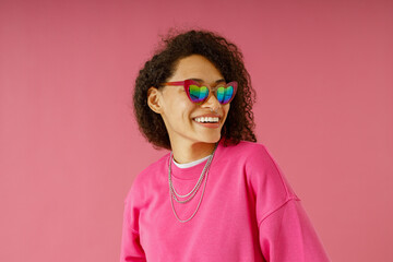 Smiling woman in sweater and heart-shaped glasses looking at side on pink studio background