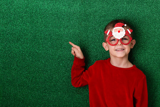 a cute boy in New Year's souvenir glasses with Santa Claus on a green background points to the background. New Year - Powered by Adobe