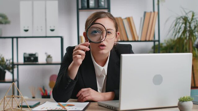 Investigator researcher scientist business woman working at office holding magnifying glass near face looking into camera with big zoomed funny eye, searching, analysing. Professional freelancer girl