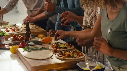Group of friends at home in kitchen adding toppings to homemade pizzas for party together - shot in slow motion
