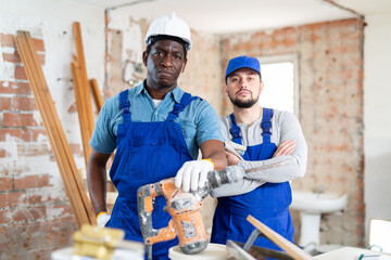Portrait of two confident builders standing at a construction site indoors with a hammer drill