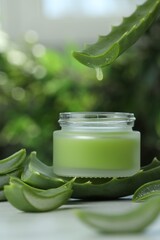 Aloe vera juice dripping from leaf into jar with cream on white table against blurred background
