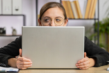 Confident business woman hiding behind laptop computer, looking at camera, spying his colleagues...