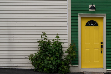 The exterior wall of a white and green house with a vibrant yellow colored door. There's a small semi-circular window in the solid metal door. The wall has a black light fixture over the closed door. 