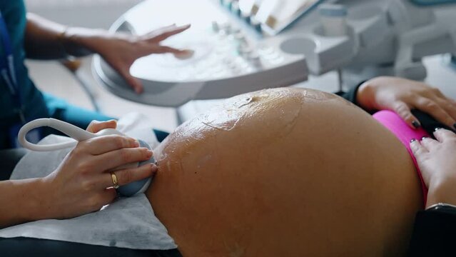 Cropped image of a pregnant lady having ultrasound check up. Hand of medic holds the device on belly and another hand operates ultrasonic machine. Close up.