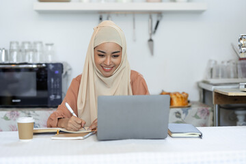 Young business woman wearing hijab sitting at kitchen table at home relaxing and working online using laptop and taking notes on vacation. lifestyle concept