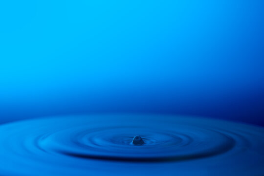 A droplet of water dispersing on a blue surface, alongside empty space for adding text. © Siradanai