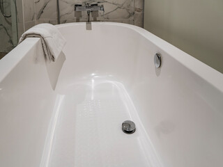 A fragment of a white oval acrylic bathtub. Shiny metal valves for draining water, taps. The towel is folded and thrown over the edge.