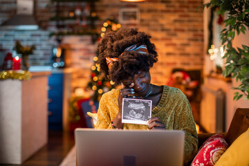 Young african american woman showing a xray of her baby showing that she is pregnant over a video call on her laptop during christmas and the new year holidays