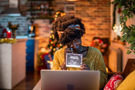 Young african american woman showing a xray of her baby showing that she is pregnant over a video call on her laptop during christmas and the new year holidays