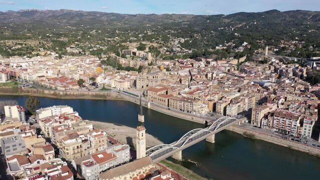 Scenic view from drone of Tortosa cityscape on banks of Ebro river with State Bridge and Ebro Battle Monument in water on sunny spring day, Spain