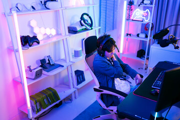 A young Asian man professional gamer wearing a jeans jacket sits on a chair and rubs a sore shoulder that suffering from stress playing a game or competition. Concept of pain from gaming and esports.