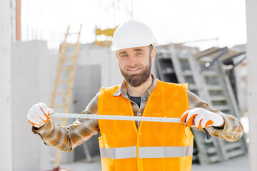 Builder repairman, foreman in protective helmet and vest stands at workplace in building and holds...