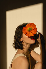 Woman hiding face by red poppy flower over neutral tan beige wall with sun light shades. Elegant...