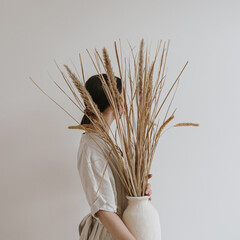 Young beautiful woman in neutral pastel beige linen dress holding sandy earthenware with dried pampas grass. Aesthetic stylish fashion tan beige coloured composition