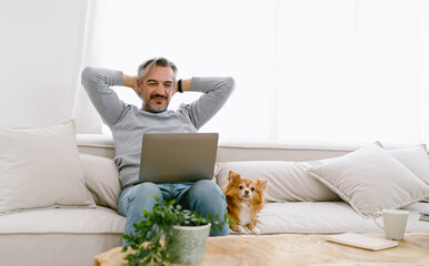 Portrait of Middle aged Caucasian man relax sitting on sofa and watching a movie on laptop with his chihuahua dog lying besides at home.  Lifestyle, Technology, domestic life, Pet friendly Concept