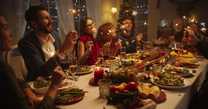 Parents, Children and Friends Enjoying Christmas Dinner Together in a Cozy Home in the Evening. Relatives Sharing Meals, Singing Traditional Festive Songs and Burning Sparkles. Slow Motion Footage