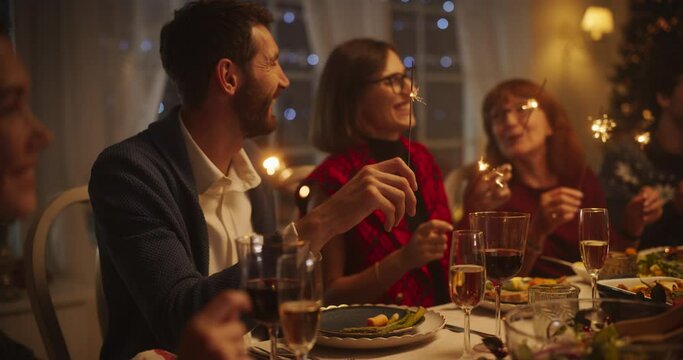 Portrait with Parents, Children and Friends Enjoying Christmas Dinner Together in a Cozy Home in the Evening. Relatives Sharing Meals, Singing Traditional Festive Songs and Burning Sparkles