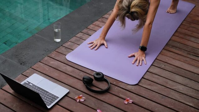 From above unrecognizable lady with laptop in sportswear concentrating and lying in balasana position on sport mat at poolside