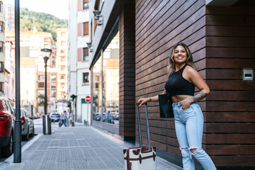 A young, joyful Latin woman in urban attire exits a city hotel, beaming with a small travel trolley.