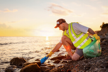 Side view of adult Caucasian bearded man volunteer wearing vest and rubber gloves picking up plastic bottle from seashore. Concept of Earth Day and environment disaster - Powered by Adobe