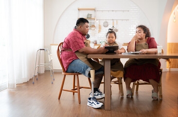 A plus-size family with a father wearing a prosthetic leg, is happily assisting a child with her homework and having fun together in the dining room of the house before cooking together