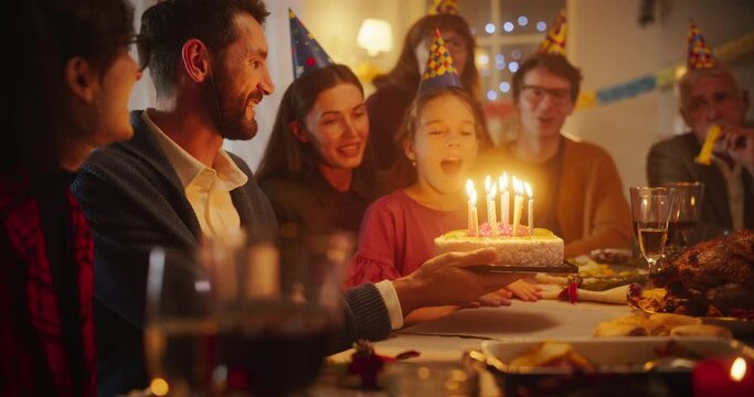 Happy Smiling Girl Blowing Out Candles on Her Delicious Birthday Cake. Young Happy Child is Surrounded by Her Family, Relatives and Friends, Sitting Behind a Table in a Cozy Living Room