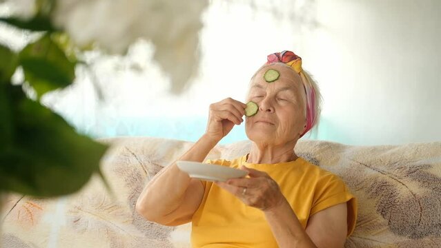 An elderly woman is sitting at home on the sofa in the living room and puts cucumber slices on her face. Anti-aging skin care, natural cosmetics.