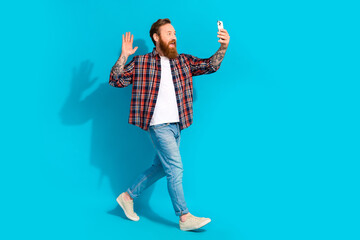 Full size photo of friendly positive man wear plaid shirt say hi on smartphone video call go empty space isolated on blue color background