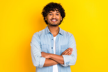 Photo of toothy beaming successful man with curly hairdo dressed denim shirt holding arms crossed isolated on yellow color background