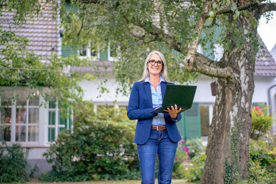 Smiling real estate agent standing with laptop in front of house