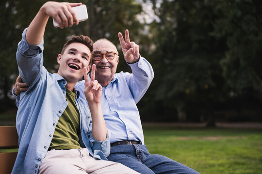Senior man and grandson sitting together on a park bench taking selfie with smartphone