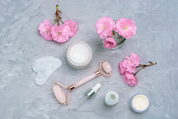 Cream jar, face roller, gua sha stone, serum bottle with pipette and pink cherry blossom on grey...