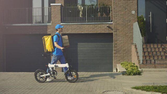 Side View of the Handsome Bike Courier Walking Holding Paper Boxes, Wearing Blue Uniform, Yellow Backpack And Helmet. Bicycle Delivery Guy Going to the Clients