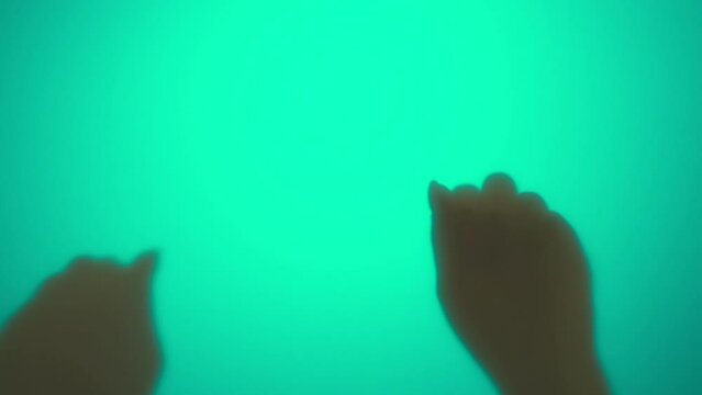 Video of fists of unrecognizable woman knocking on surface of green wall illuminated by contour light, stopping and putting open palms to background, conveying horrible scene at Halloween night.