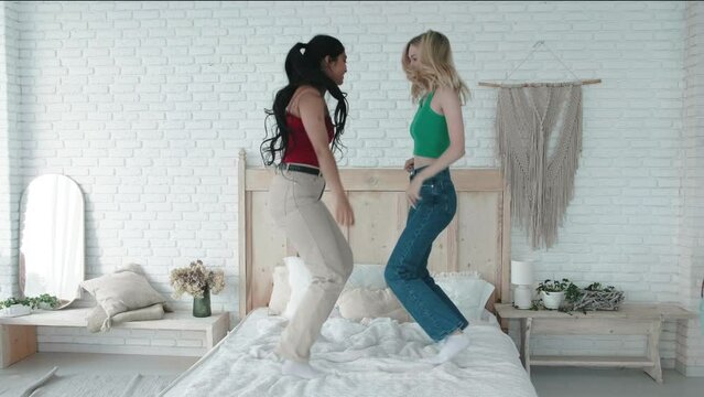 Happy joyful two females girls friends jumping on bed together, having fun, singing and dancing on the bed. Carefree positive young mixed race women. Happy friendship