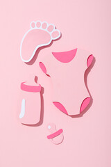 Paper baby clothes and pacifiers on pink background, top view