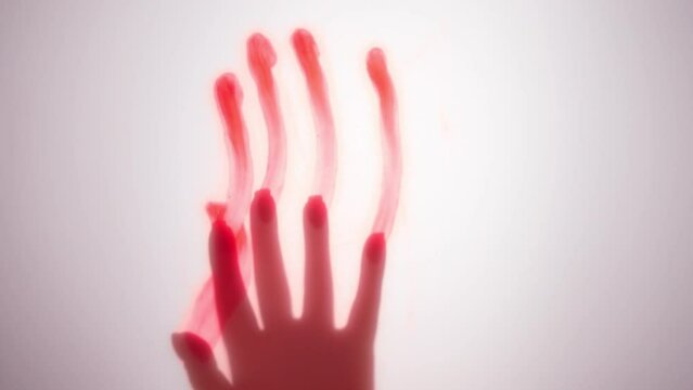 Video of hand of unrecognizable woman, smeared with red paint, touching white wall illuminated by contour light and sliding down, leaving bloody handprint on background, creating art for Halloween.